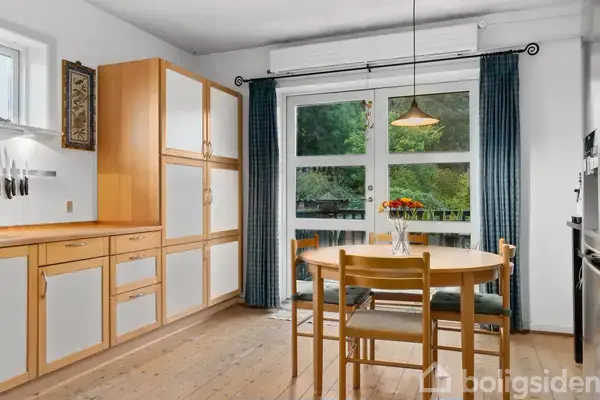Et spisebord med fire stole står ved et stort vindue med udsigt til en terrasse. Et køleskab og en kaffemaskine er placeret mod en væg med åbne hylder.