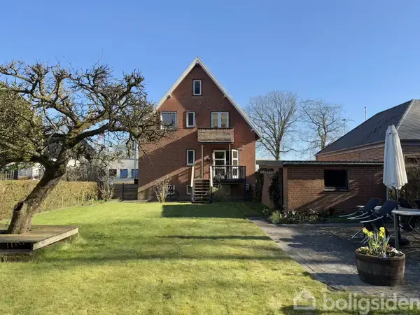 Soveværelse med dobbeltseng dækket af hvidt sengetæppe, ved siden af et skrivebord og stol. Et vindue bagved en lænestol med udsigt til træer og himmel. På gulvet ligger parket.