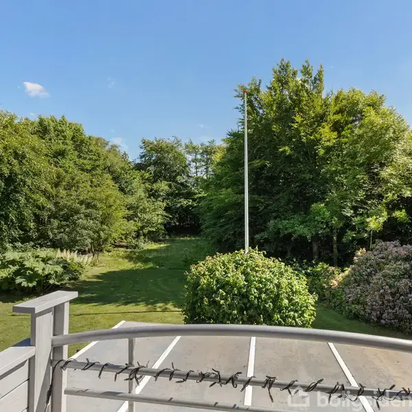 Balkon med hvidt rækværk, der vender mod en frodig have. Græsplæne og buske omgiver flagstangen i midten, under en klar blå himmel.
