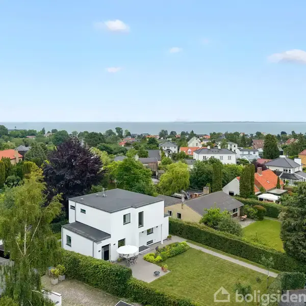Huse ses fra oven i en forstad med haver, træer og veje. Havet ligger i baggrunden, hvilket skaber en fredelig atmosfære.