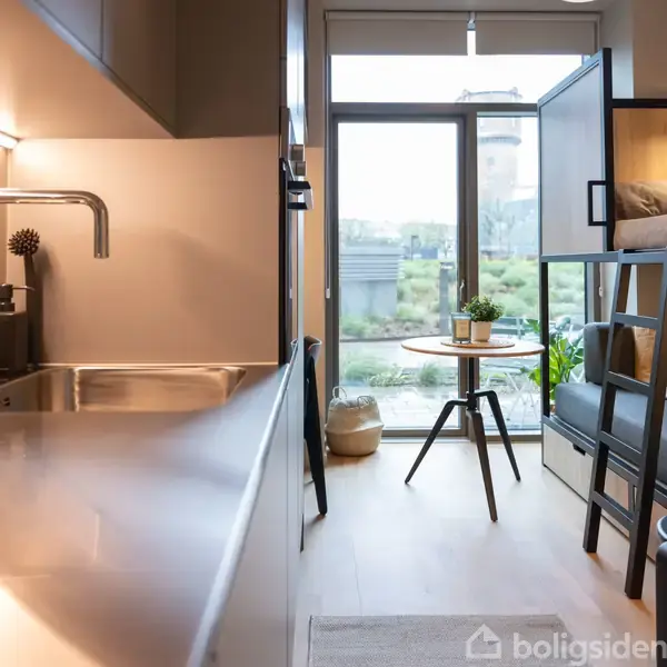 Et kompakt køkken med vask og blank bordplade, der grænser op til en funktionel stue med køjeseng og sofa. Et bord står ved et stort vindue med udsigt til grønt område.