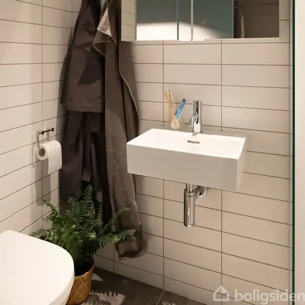 Hvid håndvask hænger på væg med hvide fliser, tandbørster ved siden af. Håndklæder hænger ved siden af toiletrulleholder. En lille grøn plante står ved væggen.