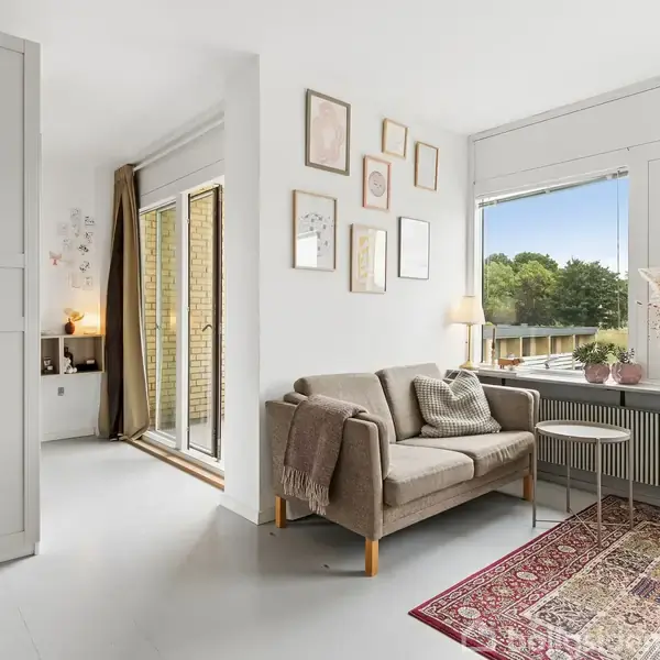 Sofa i stue med tæppe og sofabord, omgivet af vægdekorationer og store vinduer med udsigt til grønne træer. En dør fører til en tilstødende balkon.