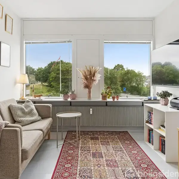 Sofa står i stue med tæppe og sofabord. Store vinduer viser udsigt over grønne træer. På væggen hænger billeder, og der står en lampe ved siden af sofaen.