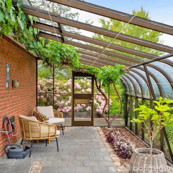 En overdækket terrasse med gennemsigtigt buet tag og klatreplanter. To stole og en plante i kurv står på stengulv. Omkringliggende have med blomstrende buske og græs.