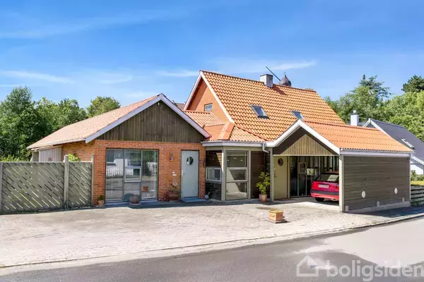 Et rødt murstenshus med rødt tegltag står stille, omgivet af en gårdsplads med en parkering. En rød bil er parkeret i en carport. Huset er omkranset af grønne træer.