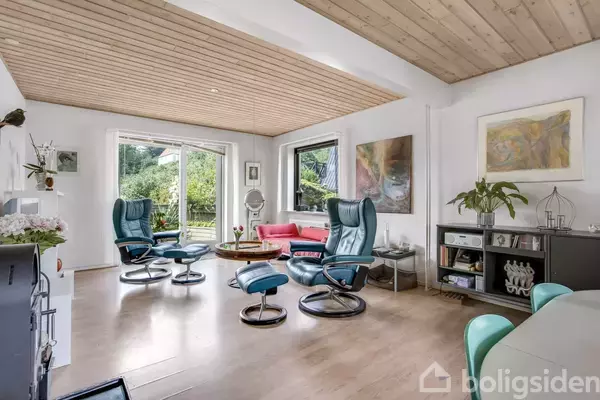 Blå lænestole står i stuen med trægulv, et stort vindue bagved åbner op til en grøn have. Malerier hænger på væggen, mens bogreol og sofa supplerer rummet.