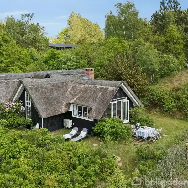 Det stråtækte hus står omgivet af frodig vegetation, med havemøbler placeret foran og på græsarealet. Skoven i baggrunden skaber en fredelig og naturlig atmosfære.