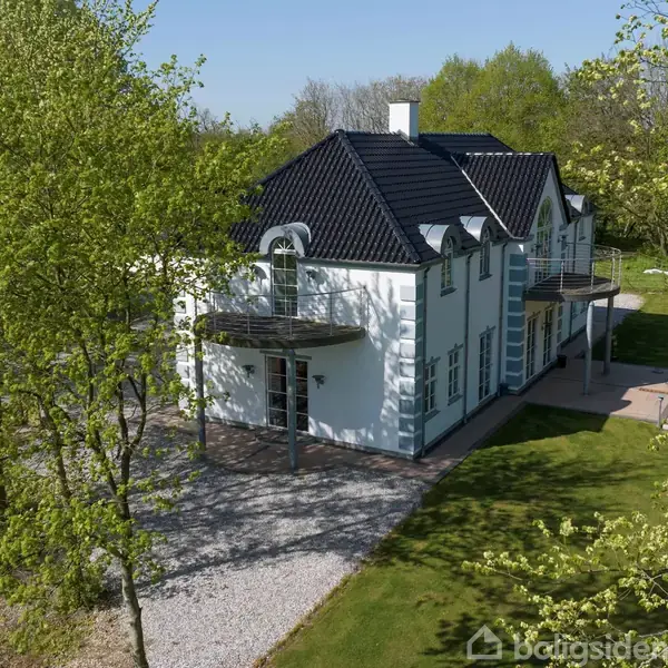 Stort, hvidt hus med sort tag ligger fredeligt omgivet af grønne træer og græsplæner. Grusvej fører op til hovedindgangen.