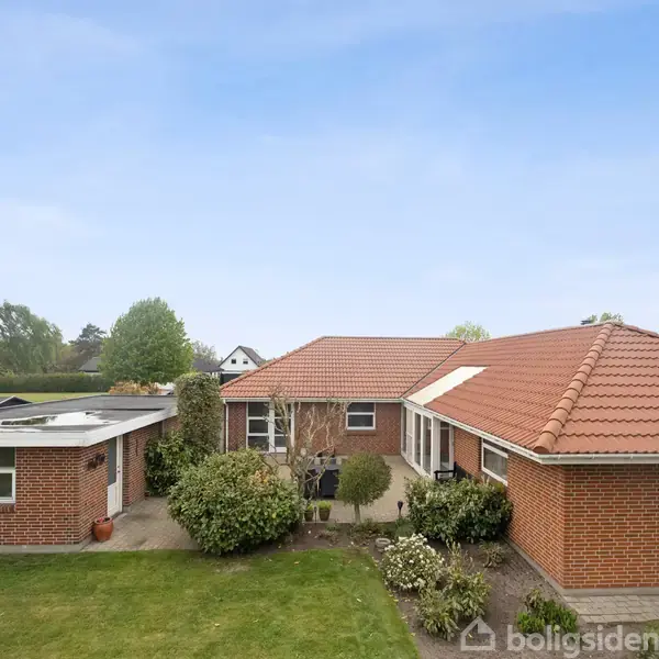 Et rødt murstenshus med rødt tegltag står i en have med græsplæne og buske. Huset er placeret i et kvarter med flere andre huse synlige i baggrunden.