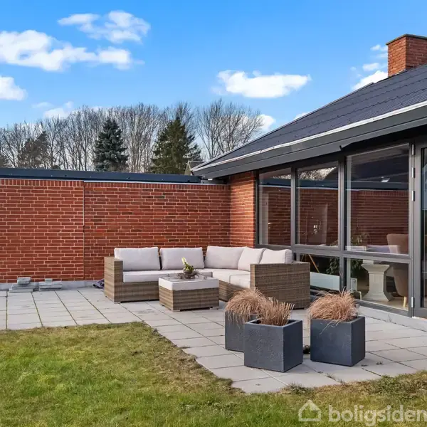 Terrasse med en grå sofa og plantekasser står ved en rød murstensbygning med store vinduer. Placeret i en have med græsplæne og træer i baggrunden under en blå himmel.