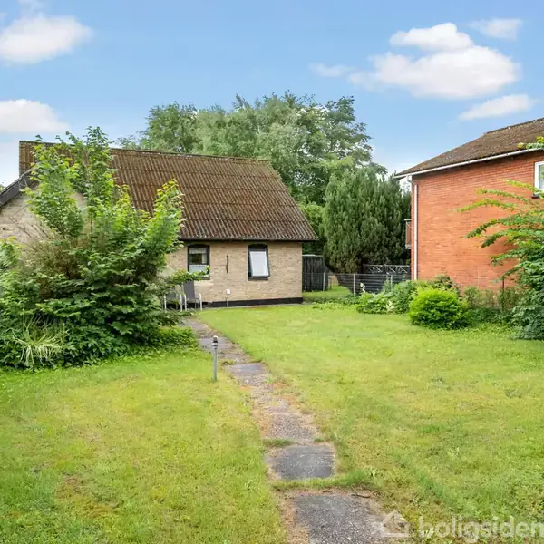 Et murstenshus står foran en grøn have med græs og buske. Til højre ses et større hus. Himlen er blå med spredte skyer.