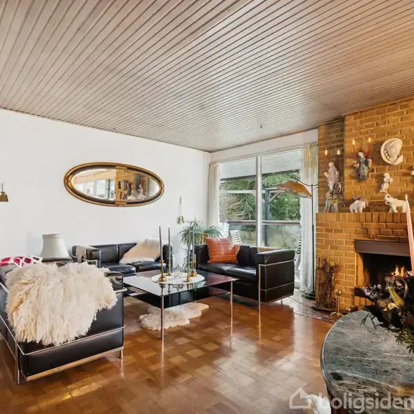 Stue med sorte lædersofaer arrangeret omkring et sofabord. Murstenspejs med dekorative figurer ved siden af vindue med udsigt til have, omgivet af trægulv og træloft.