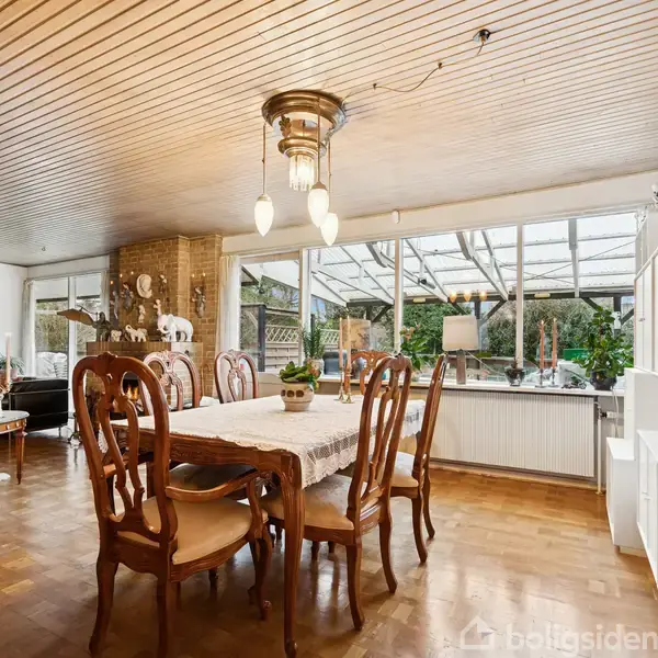 Spisebord omgivet af træstole står på parketgulv. I baggrunden ses en glasvæg med udsigt til en have. En sofa og dekorationer præger rummet, som har et træloft.