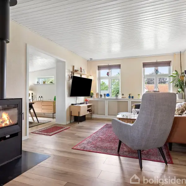 Brændeovn står tændt i en moderne stue med trægulv, store vinduer og en sofa. Et fjernsyn hænger på væggen over en reol, og rummet har et stilfuldt, hyggeligt indtryk.