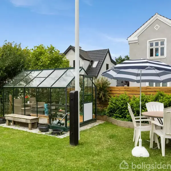 Væksthus i en grøn have ved siden af et rundt hvidt havebord med fire stole og en blå-hvid parasol. I baggrunden ses et gråt hus med rødt tag.