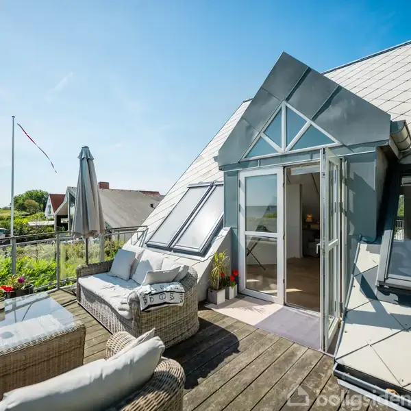 Objekt: Terrasse med flettede møbler og puder.
Handling: Møblerne står indrettet til komfortabel siddeplads.
Kontekst: Placering ved en skrånende tagflade med åbne glasdøre til huset og udsigt til en grøn have og flagstang.