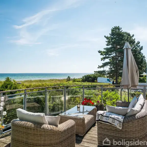To rattanmøbler med hynder står på en terrasse med glasrækværk. De vender mod havet, omgivet af grønne buske og træer. En flagstang med Dannebrog ses til højre.