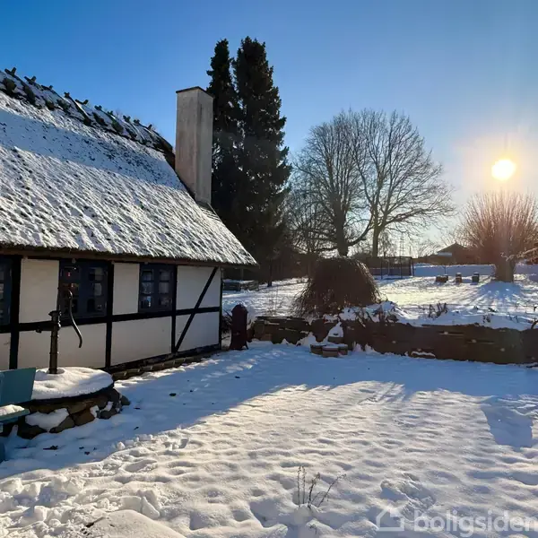 Et bindingsværkshus med stråtag står i en snedækket gård, hvor solen skinner gennem træerne i baggrunden. Der er ingen tekst i billedet.