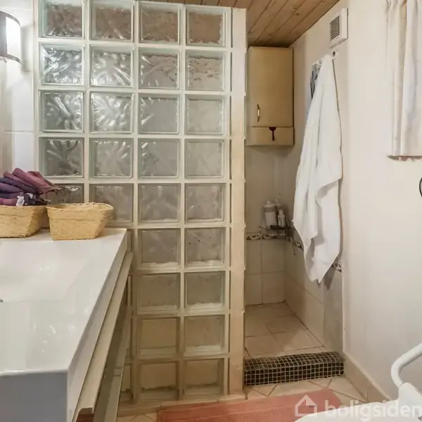 Et badeværelse med glasbyggesten som bruserafskærmning, en bred hvid vask med kurve, og håndklæder på en hylde. Håndklædestang ved vinduet, og en trælåge øverst.