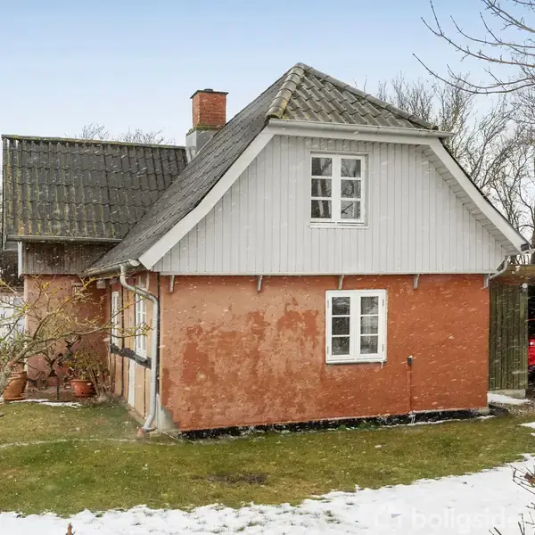 Et lille hus med rød murstensfacade og skråt tag står i en snedækket have. Et rødt køretøj er delvist synligt under en træcarport til højre.