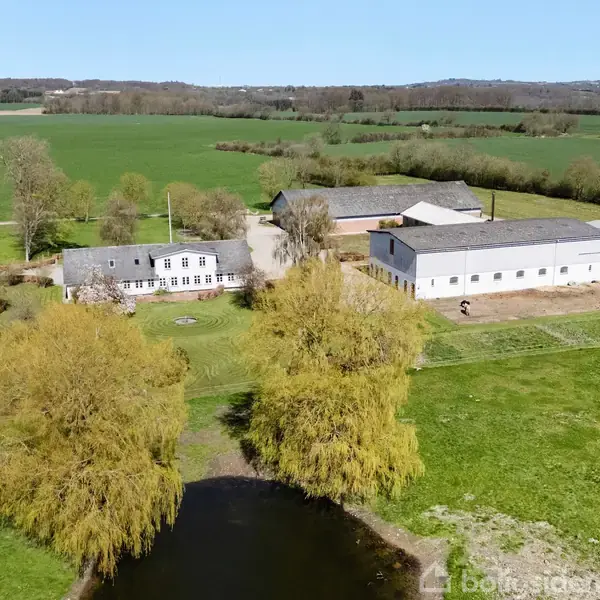 Et landligt område med en gård omringet af marker. En stor, hvid bygning og flere mindre bygninger ses nær en lille sø med træer omkring.