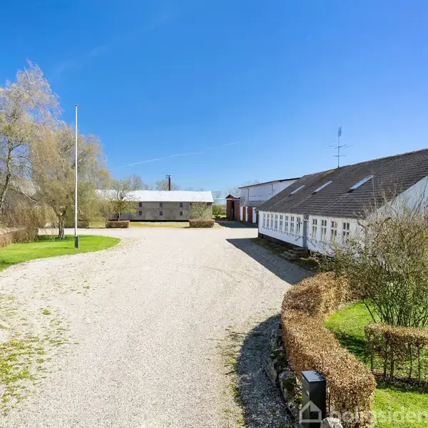 Hvide bygninger med skråtag står langs en bred grusvej. Omkring er der velplejede græsplæner og lave hække i et åbent landdistrikt med klar blå himmel.