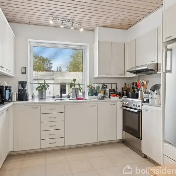 Køkken med hvide skabe og apparater, indrettet i vinkelform, indeholder kogeplader og vask. Gennem et stort vindue ses træer udenfor. Raffineret belysning og organiserede køkkenredskaber på bordpladen.