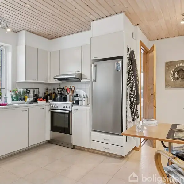 Et moderne køkken med hvide skabe og skinnende rustfrit stål apparater. En kogeplade og ovn er centralt placeret, mens et lille spisebord og stole er til højre.