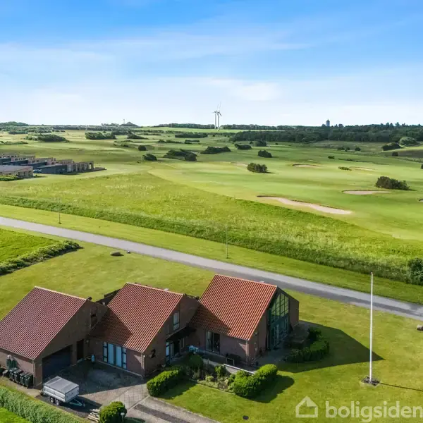 Et rødt teglhustag med tre sektioner ligger ved siden af en vej. Det er omgivet af velplejede græsplæner og en golfbane i det åbne landskab.