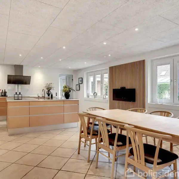 Køkken-alrum med moderne køkkenø udstyret med sort emhætte, tilstødende spisebord med træstole, store vinduer, beige flisegulv og dekorative elementer som planter og billeder på væggene.
