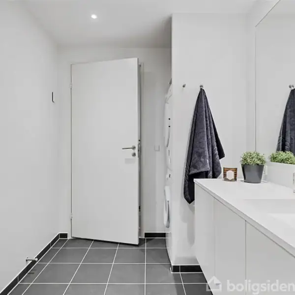 Et moderne badeværelse med en åben dør. Håndklæder hænger på væggen ved siden af en bred hvid vask. Små planter pynter på vaskebordet. Atmosfæren er minimalistisk.
