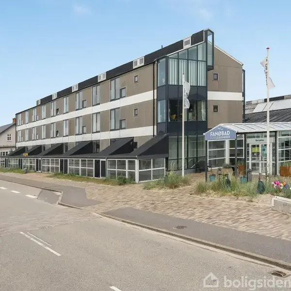 En moderne treetagers bygning med glaspartier står langs en vej. "Fanø Bad" skiltet ses ved indgangen. Omgivet af fliser og græs, med danske flag vejende i baggrunden.