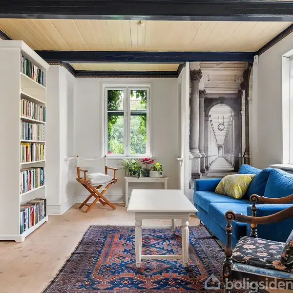 Et lyst stueområde har en blå sofa med farverige puder, en hvid reol fyldt med bøger, og et lille bord foran. Rummet er dekoreret med malerier og vinduer med udsigt til naturen.