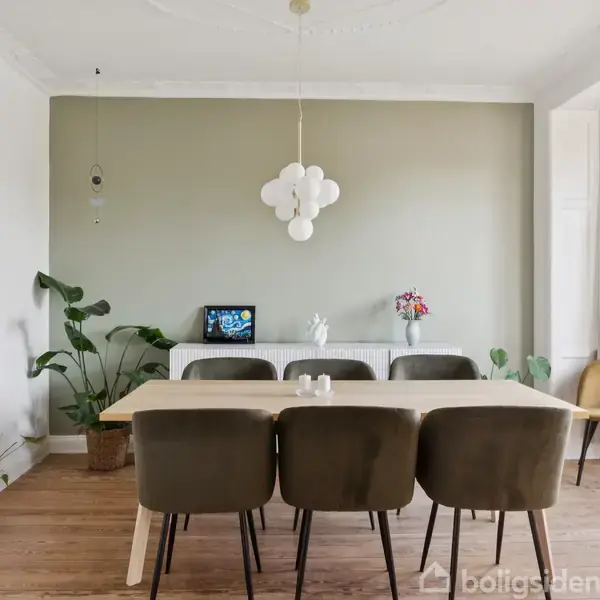 Et spisebord med seks stole står i et lyst rum med trægulv. En moderne lampe hænger over bordet. Grønne planter og kunstværker dekorerer rummets vægge og vindueskarm.