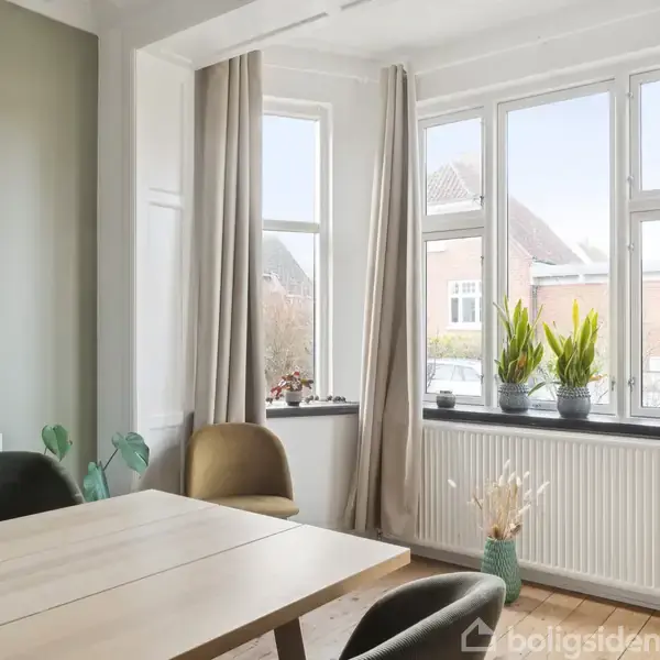 Et spisebord med to stole står ved store vinduer med beige gardiner. Planter pryder vindueskarmen, og en buket blomster står på et skab til venstre. En bygningsudsigt ses udenfor.