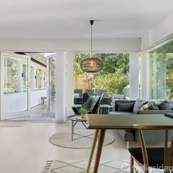 En stue med moderne møblering. Et spisebord med stole står centralt, mens en stor vinduesrude åbner op mod en terrasse med sofa og grøn have. Planter pryder rummet.