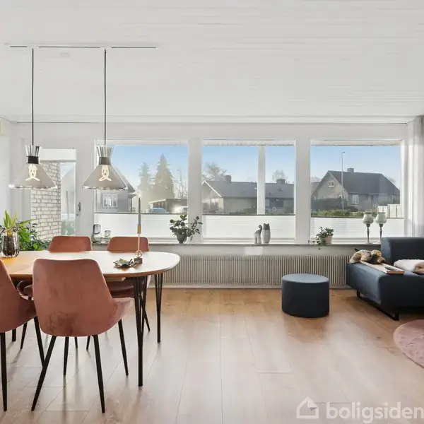 Rundt spisebord med lyserøde stole står under loftlamper. Til højre ses en mørkegrå sofa med puder og et lille bord. Store vinduer lukker lyset ind i stuen.