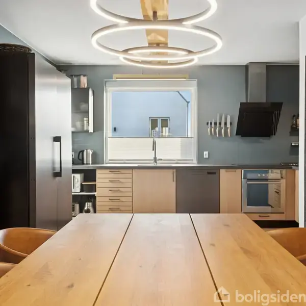 Et moderne køkken med træbord og læderstole ligger centralt. Over bordet hænger dekorative lamper. Bagved er der køkkenskabe, vask, komfur og emhætte i et stilrent design.