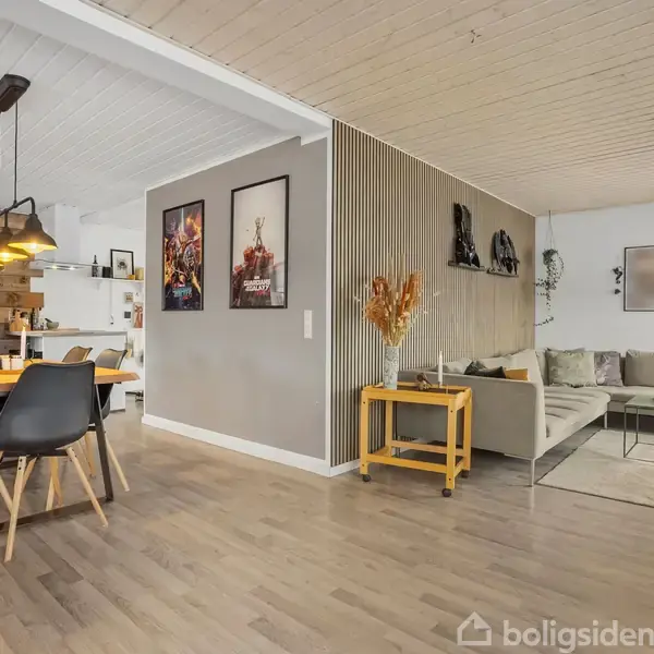 Spisestue med træbord og sorte stole. Over bordet hænger moderne lamper. Til højre en grå sofa med sofabord. Vægplakater dekorerer væggene. Rummet har lyst trægulv.