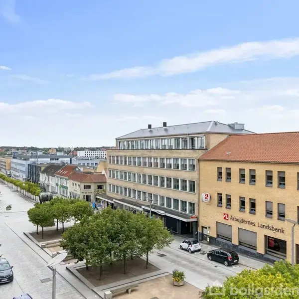 Gade med biler og træer, flankeret af bygninger, hvoraf en har skiltet "Arbejdernes Landsbank". Klar blå himmel ovenfor.