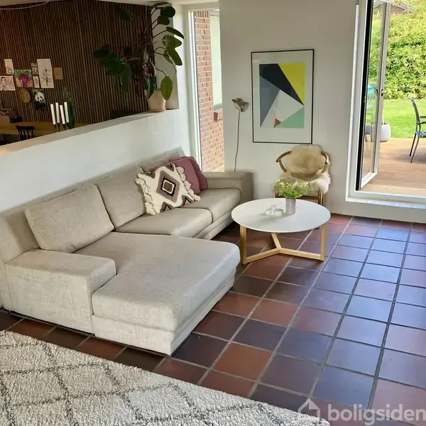 Grå sofa med pyntepuder står i en stue. Et rundt sofabord er foran sofaen. Store vinduer viser en terrasse med grønne planter udenfor.
