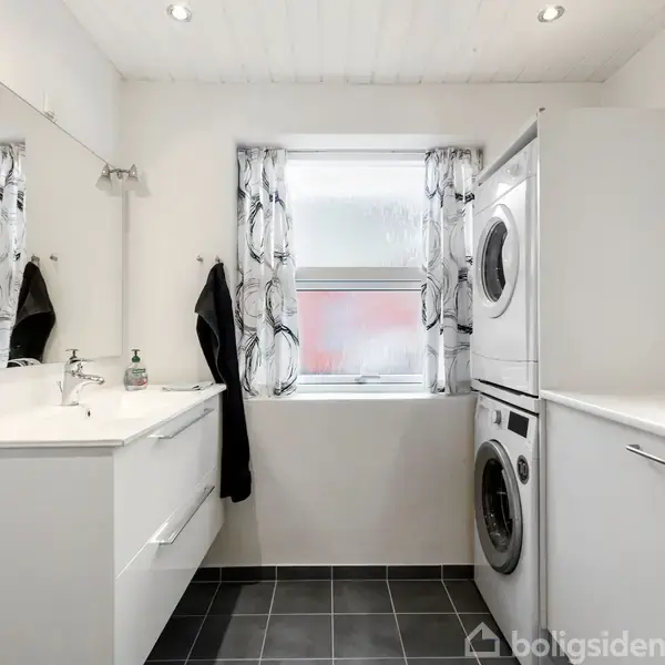 Vaskemaskine og tørretumbler er stablet oven på hinanden i et moderne badeværelse. En vask med spejl er på venstre side, og der er et vindue med gardiner.