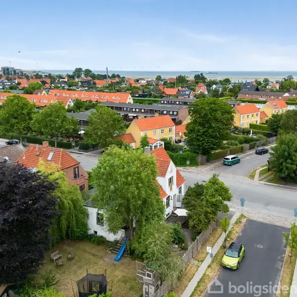 Rækkehuse med røde tage ligger i et fredeligt kvarter med træer og haver på en solrig dag. En vej med biler snor sig mellem husene, og havet kan ses i horisonten.
