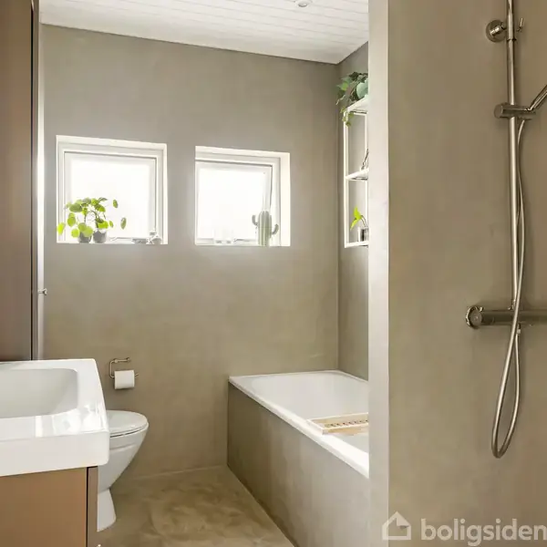 Et moderne badeværelse indeholder en vask med skab, toilet, og bruser. Badekarret er placeret ved to små vinduer med planter. Rummet har lyse vægge og gulve, som giver et rent udtryk.