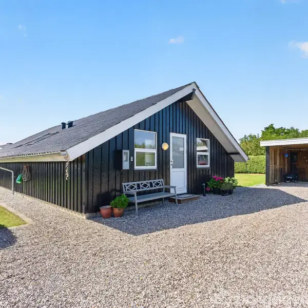 Sortmalet hus med skråt tag, stående på en grusvej. Foran huset står to potteplanter og en bænk. Til højre ses et lille skur med en overdækket terrasse.