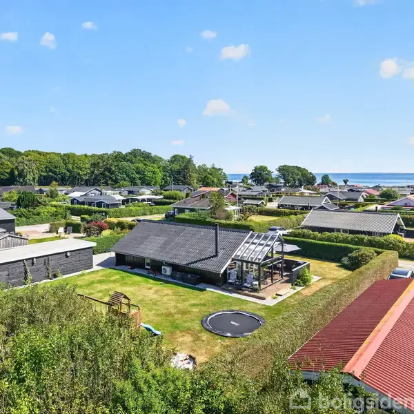 En villa med udendørs terrasse og indbygget trampolin ligger i et grønt boligområde nær havet med flere huse og træer omkring.