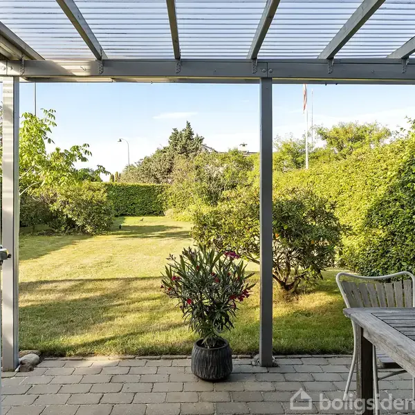 En terrasse med glastag viser et haveområde. Et bord med stole står til højre. Haven er omgivet af buske og træer under en blå himmel.