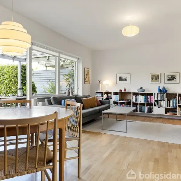 Spisestue og stue med et rundt spisebord, træstole, grå sofa, reoler med bøger og dekorationer. Store vinduer giver udsigt til en terrasse og grønne omgivelser udenfor.