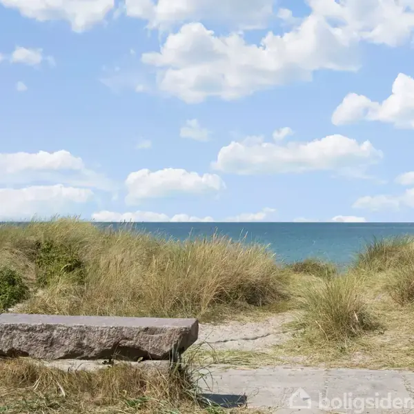 Stenbænk står stille blandt klitter med græs, vendt mod havet under en skyfuld himmel. Sand og vegetation omkranser området, mens det klare blå vand strækker sig til horisonten.